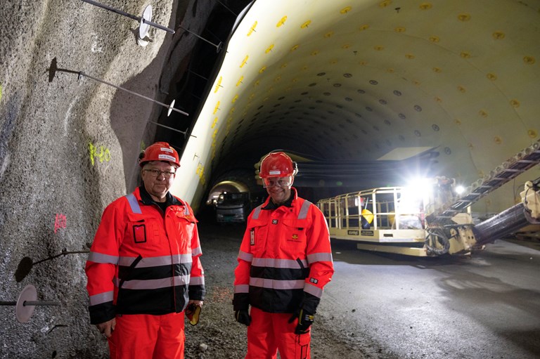Jan Bjarne Thorsnes (t.v.) og Ole Martin Lilleby frå Vegvesenet inne i tunnelen. Dei har orange vernetøy og i bakgrunnen er folk i gang med å henge opp vass- og frostsikringsmatter i tunneltaket.
