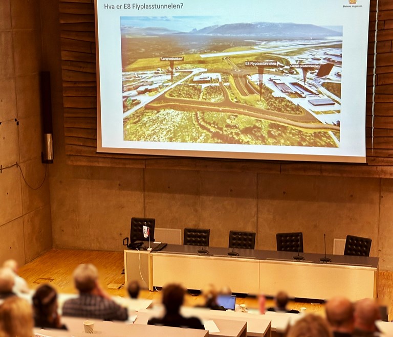 Bildet viser folk fotografert bakfra mes de sitter i kommunestyresalen i Tromsø og ser på en presentasjon av Flyplasstunnelen. Bildeteksten sier; Åpent møte for grunneiere i kommunestyresalen i Tromsø.