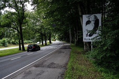 Bil som kjører gjennom skog og kampanjeskilt for å huske bilbelte til høyre for veien.