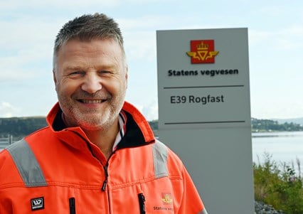 Portrettbilde av prosjektleder Oddvar Kaarmo ikledd oransje Statens vegvesen-jakke foran Rogfast sin brakkerigg. 